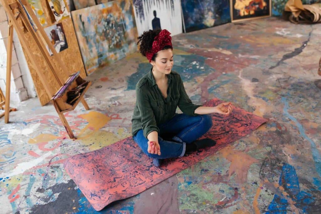 Young beautiful woman dreamily closing eyes sitting on yoga mat in lotus pose and meditating spending time in big cozy workshop with wood easel and modern paintings on background