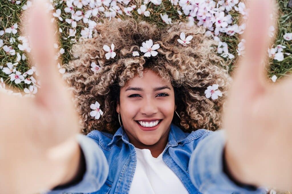 top view of happy hispanic woman with afro hair lying on grass among pink blossom flowers. Springtime
